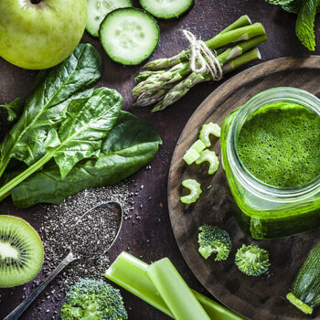 Top view of a rustic table filled with green fruits and vegetables for a perfect detox diet. At the left of an horizontal frame is a glass with fresh green smoothie and a large variety of fruits and vegetables are all around it. Predominant color is green. Low key DSRL studio photo taken with Canon EOS 5D Mk II and Canon EF 100mm f/2.8L Macro IS USM