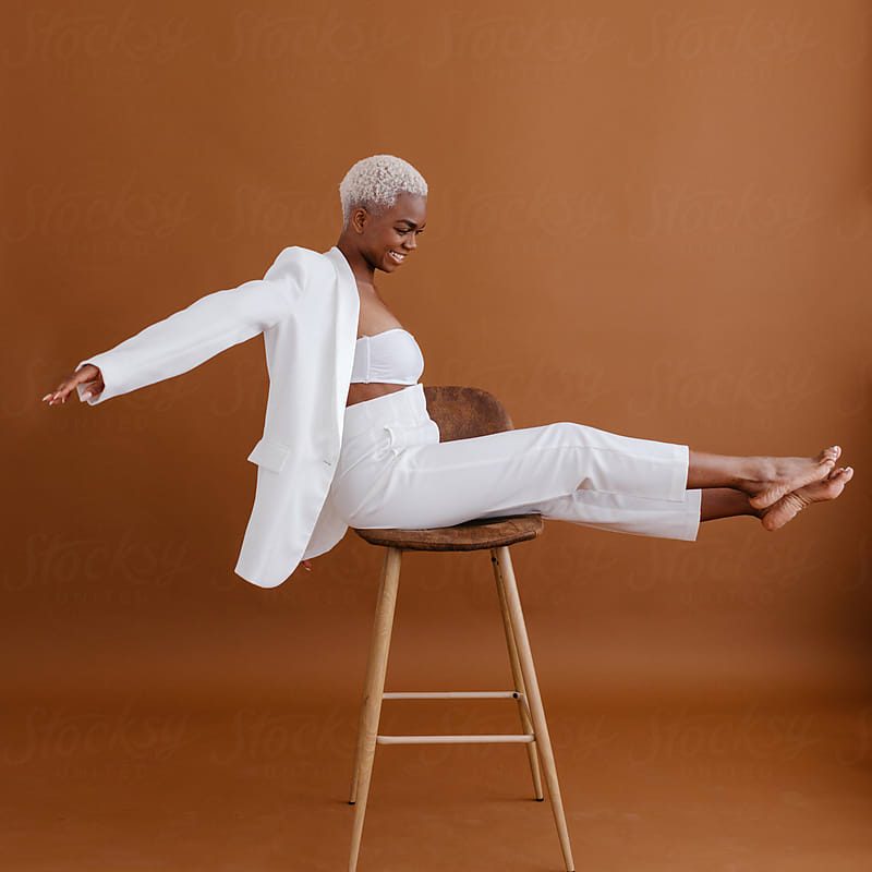 Stocksy_comp_watermarked_4893686 Side view of cheerful young African American female in white suit and bra spreading arms and smiling while balancing on chair against brown background.